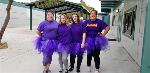 4 female all wearing purple shirts and tutus posing in front of classroom